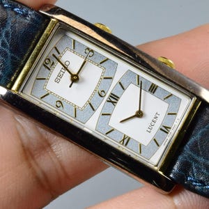 Puede incluir: Un reloj de pulsera Seiko Lucent rectangular con caja plateada y correa de cuero azul. La esfera del reloj tiene dos secciones con agujas doradas y números romanos. El reloj está sujeto por una mano.