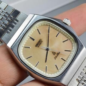 Pode incluir: Um relógio de pulso Seiko Quartz em tom prateado com caixa retangular e mostrador bege claro. O relógio apresenta marcadores de hora pretos finos, ponteiros pretos e uma pulseira de elos de metal. O texto Seiko Quartz e SilverWave está impresso.