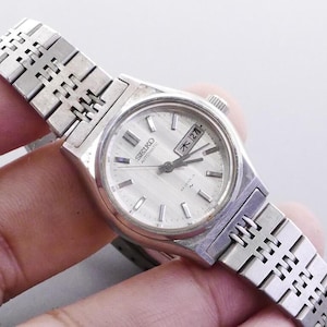 Puede incluir: Un reloj de pulsera automático Seiko vintage con esfera plateada y correa de acero inoxidable. La esfera muestra el día y la fecha. La correa tiene un diseño texturizado único. Un reloj clásico.