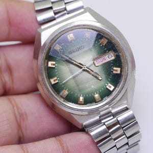 Puede incluir: Un reloj de pulsera Seiko plateado con una esfera verde y una correa de metal plateado. La esfera del reloj tiene marcadores de hora de color dorado y una ventana de fecha que dice "SUN 6".