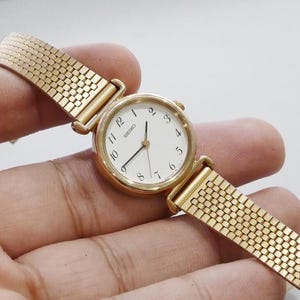 Op de afbeelding: Een goudkleurig Seiko polshorloge met een witte wijzerplaat en zwarte wijzers. Het horloge heeft een ronde wijzerplaat met zwarte cijfers en een mesh band. Het horloge wordt door een hand vastgehouden.