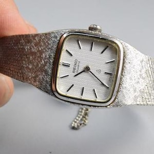 May include: A silver-tone Seiko Quartz wristwatch with a square face, white textured dial, and black hour markers. The watch features a metallic band with a cross-hatched, brushed finish and a small crown on the side for time adjustment.