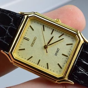 Könnte beinhalten: Eine rechteckige, goldfarbene Seiko Spirit Armbanduhr mit champagnerfarbenem Zifferblatt, dünnen goldenen Stundenmarkierungen und goldenen Zeigern. Die Uhr hat ein schwarzes Lederarmband mit Krokodilprägung und eine kleine goldene Krone.