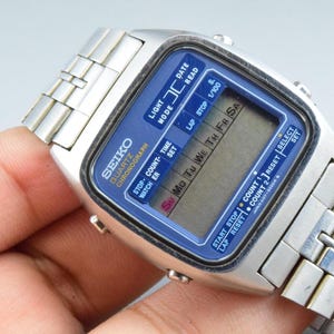 Pode incluir: Um relógio digital Seiko Quartz Chronograph de tom prateado com uma pulseira de elos metálicos. O mostrador apresenta uma borda azul com texto branco, incluindo "LIGHT", "MODE", "DATE", "READ", "STOP", "COUNT", "TIME", "SET" e "START STOP".