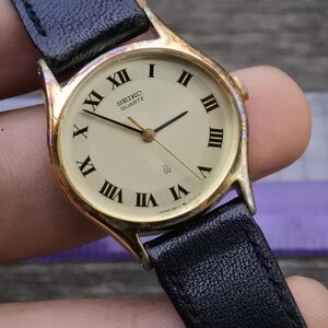 May include: A gold-toned Seiko Quartz watch with a cream-colored face and black Roman numeral hour markers. The watch has black hands and a black leather band. The watch face displays the Seiko Quartz logo.