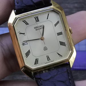 Può includere: Un orologio Seiko Quartz dorato con quadrante rettangolare e numeri romani neri. L'orologio ha un quadrante color crema, lancette nere e un cinturino in pelle nera. Stile vintage.