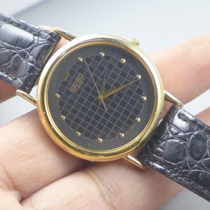 Könnte beinhalten: Eine goldfarbene Seiko Quartz-Uhr mit einem schwarzen Zifferblatt mit Gittermuster und goldfarbenen Stundenmarkierungen. Die Uhr hat goldfarbene Zeiger und ein schwarzes Lederarmband mit Krokodilmuster.