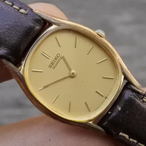 Puede incluir: Un reloj Seiko Quartz dorado con una correa de cuero marrón. La esfera del reloj es de forma ovalada y tiene un fondo dorado con marcadores de hora en números romanos negros.