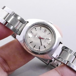 Könnte beinhalten: Eine silberfarbene Seiko Armbanduhr mit hellem Zifferblatt und Datumsanzeige auf 3 Uhr. Die Uhr hat ein silbernes Armband und einen roten Sekundenzeiger. Die Uhr ist im Vintage-Stil.