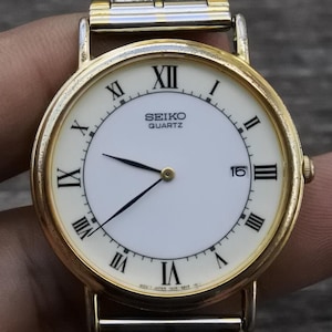 May include: A gold-tone Seiko quartz watch with Roman numeral hour markers and a white face. The watch has a two-tone band with a silver and gold finish.