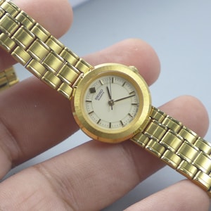 Könnte beinhalten: Eine goldfarbene Seiko Quartz Armbanduhr mit einem rechteckigen Gliederarmband. Das Zifferblatt der Uhr ist cremefarben mit schwarzen Zeigern und Markierungen. Die Uhr wird vor einem neutralen Hintergrund präsentiert.