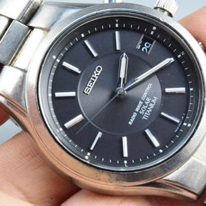 Könnte beinhalten: Eine Seiko Armbanduhr mit einem silberfarbenen Titan-Gliederarmband und einem runden schwarzen Zifferblatt. Das Zifferblatt zeigt silberne Stundenmarkierungen, silberne Zeiger, ein Datumsfenster auf der Drei-Uhr-Position und den Text Seiko, Radio Wave Control, Solar, Titanium.