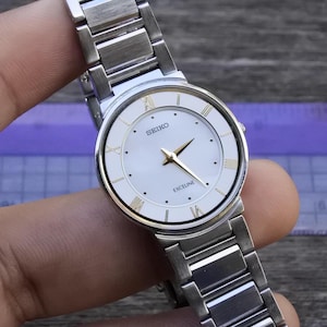 May include: A silver Seiko Exceline watch with a white face and gold hands and Roman numeral hour markers. The watch has a silver band and is held against a wooden surface.