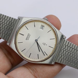 Op de afbeelding: Een vintage Seiko Quartz horloge met een zilverkleurige rechthoekige kast en een mesh band. De lichtgekleurde wijzerplaat heeft zwarte uurmarkeringen en wijzers, en een datumweergave. Een klassiek horloge, ongeveer 3,5 cm breed.