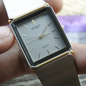 Puede incluir: Un reloj Seiko Spirit rectangular con una correa plateada y detalles dorados. La esfera es blanca con manecillas e indicadores dorados, y la palabra "SPIRIT" está impresa encima de "SEIKO".