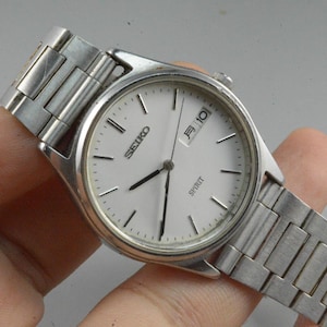 Könnte beinhalten: Eine silberne Seiko Spirit Armbanduhr mit weißem Zifferblatt, schwarzen Zeigern und Datumsanzeige. Die Uhr hat ein silbernes Armband und wird in einer Hand gehalten.