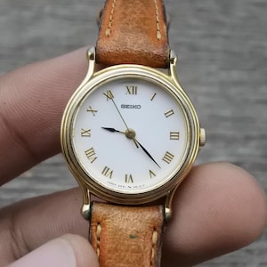 May include: A gold-tone wristwatch with a white face and Roman numeral hour markers. The watch has a brown leather band.