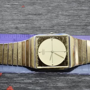 Puede incluir: Un reloj de pulsera dorado con una esfera cuadrada y una correa de metal dorado. La esfera del reloj es blanca con agujas negras y la palabra "SEIKO" y "QUARTZ" impresas en negro.