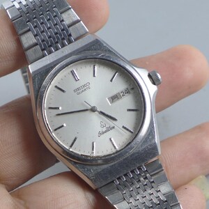 May include: Silver Seiko Quartz watch with a silver dial and a date display at the 3 o'clock position. The watch has a silver bracelet and a round face with black hour markers. The watch face also has the words "Seiko Quartz" and "SilverWave".