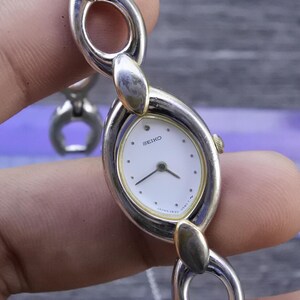 May include: A silver and gold-toned Seiko wristwatch with an oval face and a white dial. The watch has a linked bracelet and a small gold crown. The watch face displays the Seiko logo and the words "JAPAN 1820".