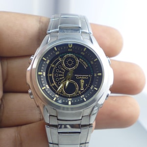May include: Silver-toned Casio watch with a black and gold face. The watch features multiple dials and the brand name "CASIO" is visible. The watch has a stainless steel band and is held by a hand.