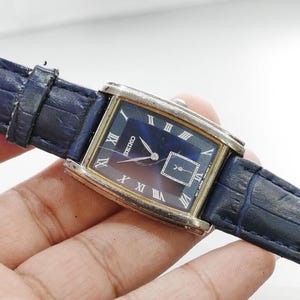 Pode incluir: Um relógio Seiko retangular com mostrador azul escuro e caixa prateada. O relógio apresenta algarismos romanos e um pequeno subdial quadrado. Possui uma pulseira de couro azul escuro.