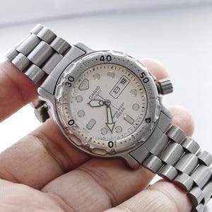 Puede incluir: Un reloj Casio Quartz plateado con esfera blanca, visualización de fecha y bisel giratorio. El reloj tiene una correa de acero inoxidable y es resistente al agua hasta 200 m. El reloj se sostiene en una mano.
