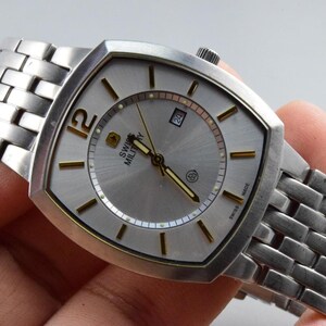 Könnte beinhalten: Eine silberne Swiss Military Uhr mit rechteckigem Zifferblatt und silbernem Armband. Das Zifferblatt hat goldfarbene Akzente und eine Datumsanzeige. Die Uhr wird von einer Hand gehalten.