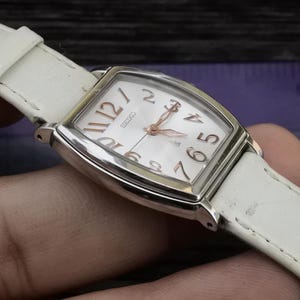 Può includere: Un orologio da polso in pelle bianca con una cassa rettangolare argentata e un quadrante bianco con numeri romani neri. Il quadrante dell'orologio ha anche lancette nere e un piccolo datario. L'orologio è al polso di una persona.