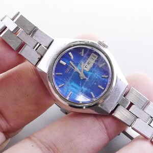 Puede incluir: Un reloj Seiko vintage con una correa de metal plateado y una esfera azul. La esfera del reloj muestra el día y la fecha. El reloj se muestra sobre un fondo claro.