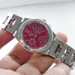 Könnte beinhalten: Eine silberfarbene Seiko-Armbanduhr mit burgunderfarbenem Zifferblatt. Die Uhr hat ein rechteckiges Gliederarmband und ein rundes Zifferblatt mit römischen Ziffern und Hilfszifferblättern. Die Uhr wird in einer Hand gehalten.