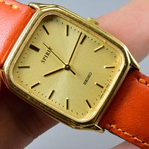 Puede incluir: Un reloj Seiko Spiria dorado con esfera cuadrada y esfera dorada. El reloj tiene marcadores de hora y agujas negros. Está sujeto a una correa de cuero naranja brillante con costuras amarillas.