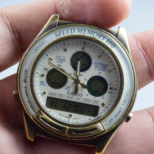 May include: A Casio Speed Memory 100 wristwatch with a gold-tone case and white face. The watch features multiple dials, digital display showing "AL 12:00 -3", and text including "SPEED MEMORY 100" and "CASIO".