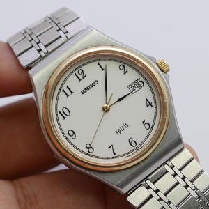 Könnte beinhalten: Eine silberfarbene Seiko Spirit-Uhr mit weißem Zifferblatt, goldfarbener Lünette und Datumsanzeige bei 3 Uhr. Die Uhr hat ein silbernes Armband und goldfarbene Akzente an Krone und Zeigern. Das Wort "SEIKO" ist oben aufgedruckt.