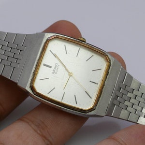 Puede incluir: Un reloj de pulsera Seiko de cuarzo plateado y dorado con una esfera cuadrada y una pulsera de metal plateado.