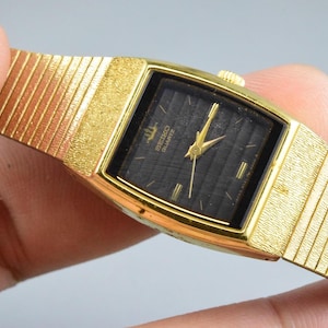 Könnte beinhalten: Eine goldfarbene Seiko Quartz Armbanduhr mit einem rechteckigen schwarzen Zifferblatt mit horizontalen, strukturierten Linien, goldfarbenen Zeigern und einem passenden goldfarbenen Metallgliederarmband mit strukturierter Oberfläche am Gehäuserand.
