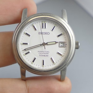 May include: A silver Seiko Perpetual Calendar Titanium watch face with a white dial. The watch has black hour markers, a date window at the 3 o'clock position, and the Seiko logo. The watch is held by a hand.