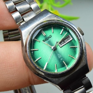 Op de afbeelding: Vintage Seiko Quartz horloge met een zilverkleurige kast en band. De wijzerplaat is heldergroen met zilveren uurmarkeringen en wijzers. De dag en datum worden weergegeven in een klein venster. Een klassiek horloge.