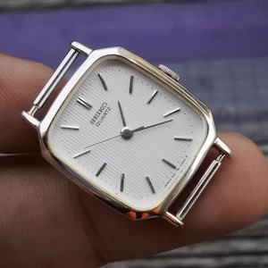 Puede incluir: Un reloj de pulsera Seiko Quartz de forma cuadrada plateado y dorado con una esfera blanca y agujas negras.