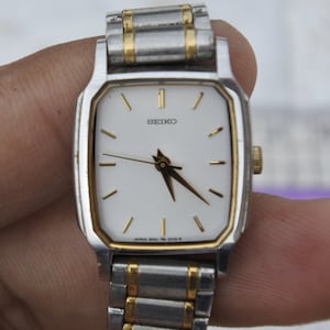 Könnte beinhalten: Eine Seiko-Uhr mit weißem Zifferblatt, goldfarbenen Zeigern und einem silber-goldfarbenen Armband. Die Uhr hat ein rechteckiges Zifferblatt mit goldenen Akzenten und ein silbernes Armband mit goldfarbenen Gliedern. Das Zifferblatt zeigt die Marke "SEIKO".