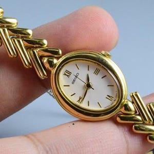 May include: A gold-tone Seiko wristwatch featuring an oval-shaped case with a white dial, black Roman numeral hour markers, and slender hands. The watch is attached to a flexible, textured gold-tone metal link bracelet with a repeating chevron pattern.