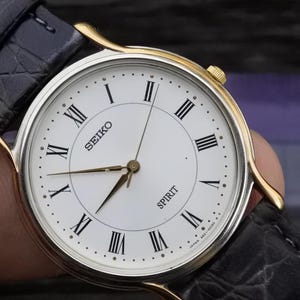 May include: A gold-tone Seiko Spirit watch with a white face and black Roman numeral hour markers. The watch has a black leather band with a crocodile pattern.