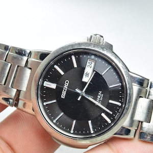 May include: A silver-toned Seiko wristwatch with a black circular dial, silver hour markers, and a metallic link bracelet. The watch face displays the brand name Seiko, the word Titanium, 10 BAR, and a day-date window showing the character 金 and 24.