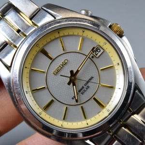 Könnte beinhalten: Eine silber- und goldfarbene Seiko Armbanduhr mit hellgrauem Zifferblatt. Die Uhr hat goldfarbene Zeiger und Stundenmarkierungen, eine Datumsanzeige bei 3 Uhr und den Text "SEIKO" und "RADIO WAVE CONTROL SOLAR". Das Armband ist silberfarben.