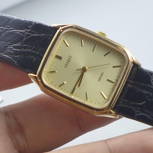Könnte beinhalten: Eine goldfarbene Seiko Spirit Armbanduhr mit quadratischem Zifferblatt und schwarzem Lederarmband. Das Zifferblatt ist hellgoldfarben mit schwarzen Stundenmarkierungen und Zeigern. Das Wort "Spirit" ist auf dem Zifferblatt aufgedruckt.