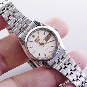 Puede incluir: Un reloj de pulsera automático Seiko plateado con esfera blanca y agujas e indicadores dorados. El reloj presenta una pantalla de día y fecha y una pulsera de eslabones plateados. El reloj está sujeto por una mano.