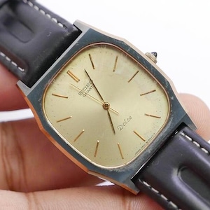 Puede incluir: Un reloj Seiko Quartz Dolce dorado con una esfera cuadrada y una correa de cuero negro. La esfera del reloj tiene agujas y marcadores dorados, y la palabra "Dolce" está impresa en dorado debajo del centro.