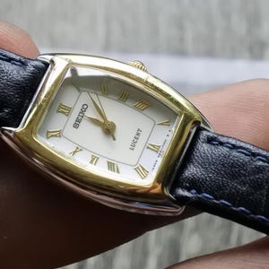 May include: A gold and silver women's wristwatch with a white face and black leather band. The watch face has gold Roman numeral hour markers and the words "SEIKO" and "LUCENT" printed on it.