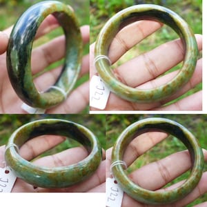 May include: A jade bangle bracelet with a mix of green, brown, and black hues. The smooth, polished surface reflects light, showcasing the natural variations in color. The bracelet is a circular shape, designed to be worn on the wrist. The tag reads "J22".