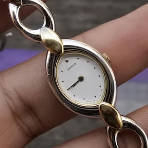 May include: A silver and gold women's wristwatch with a white face and black hands. The watch has a unique link bracelet design.